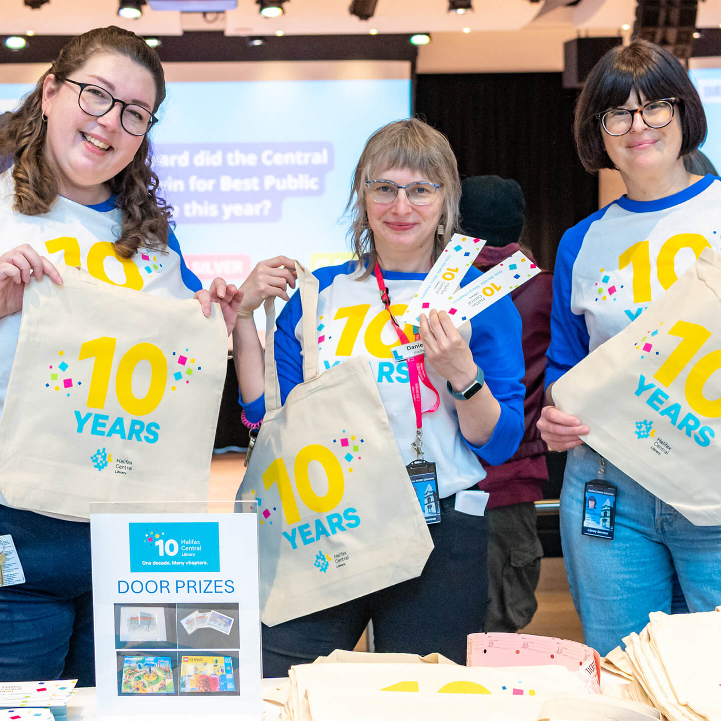 Staff holding 10 year anniversary tote bags while wearing matching anniversary shirts, smiling to camera.